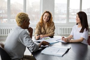 A group of people are collaborating around a desk in an office building, sitting and conversing while a person looks out the window.
