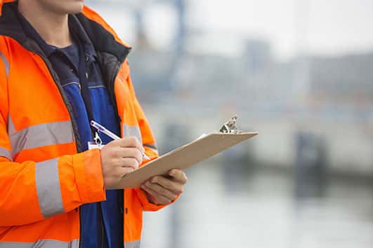 The person is standing outdoors wearing a high-visibility workwear jacket, an orange life jacket, and holding an orange.