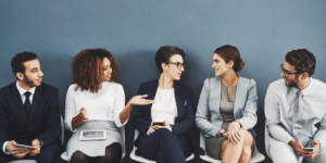 A diverse group of professionals in business attire sitting in a row, smiling and conversing superannuation while waiting, representing modern workplace trends and HR strategies.