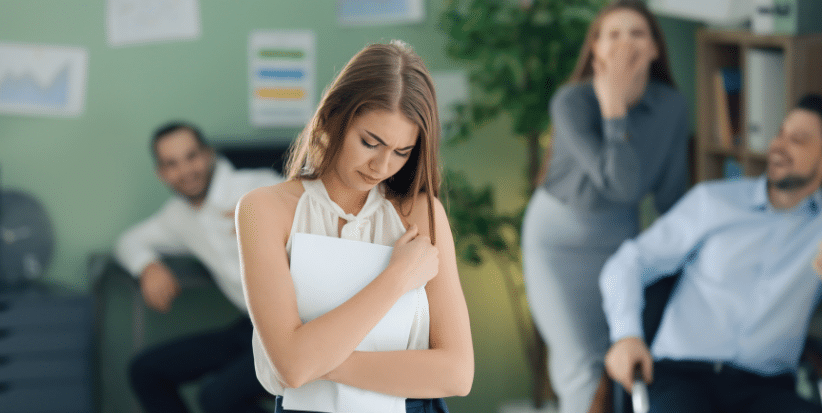 A distressed employee sitting apart from colleagues, highlighting the impact of workplace bullying and the need for safe, respectful environments.